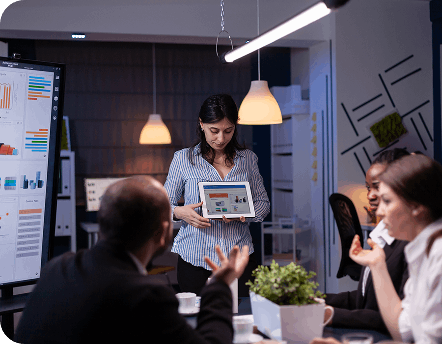 A woman stands holding a tablet with graphs displayed, presenting to three colleagues seated around a table in a modern office during a meeting. A large screen with charts is also visible.