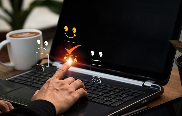 A hand touches a laptop screen, selecting a happy face with five stars for feedback. Two other faces, sad and neutral with fewer stars, are visible. A coffee cup sits beside the laptop. A hand touches a laptop screen, selecting a happy face with five stars for feedback. Two other faces, sad and neutral with fewer stars, are visible. A coffee cup sits beside the laptop.