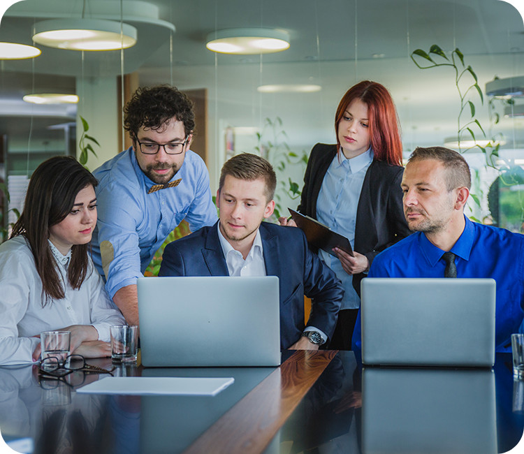Five people in business attire are gathered around a table with two laptops, having a discussion in a modern office with glass walls and plants in the background. Five people in business attire are gathered around a table with two laptops, having a discussion in a modern office with glass walls and plants in the background.