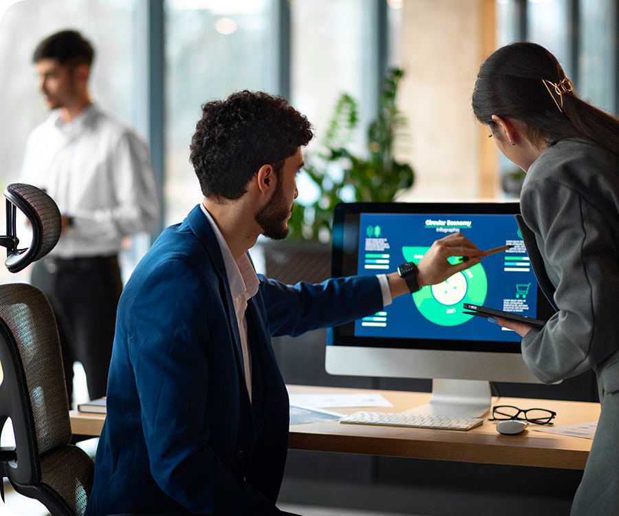 Two people in business attire discuss a circular economy infographic displayed on a computer monitor in an office, while another person stands in the background near large windows. Two people in business attire discuss a circular economy infographic displayed on a computer monitor in an office, while another person stands in the background near large windows.