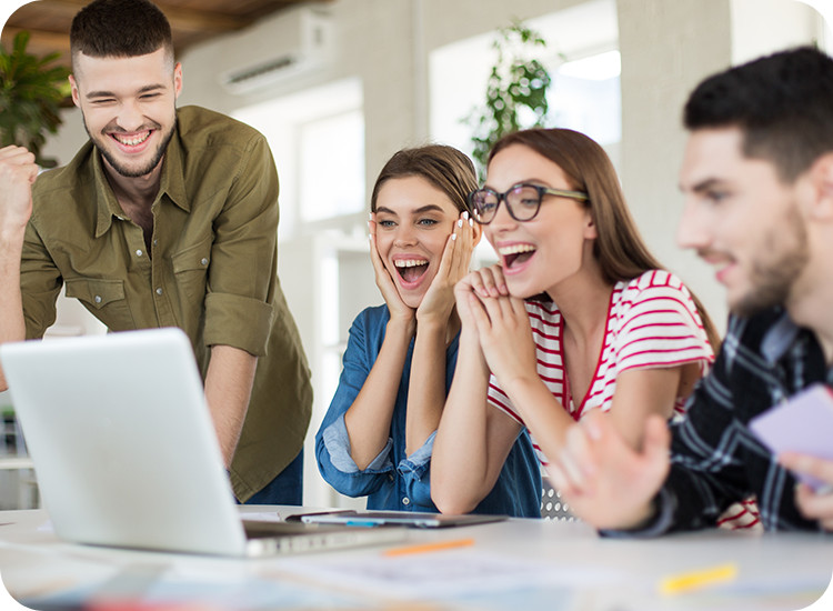 Four young adults sit around a laptop, smiling and cheering with excitement, suggesting they have received good news or achieved a goal together in a bright, modern office space. Four young adults sit around a laptop, smiling and cheering with excitement, suggesting they have received good news or achieved a goal together in a bright, modern office space.
